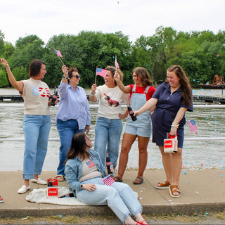 Group of people by a lake, waving American flags and enjoying a sunny day.
