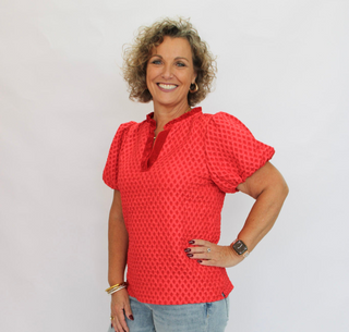 A smiling woman with curly hair stands against a white studio background wearing a bright red short-sleeve top with a textured pattern, light-wash blue jeans, and brown shoes.