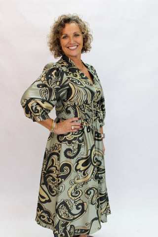 A smiling woman with curly brown hair models a long-sleeved midi dress featuring a black, beige, and olive green abstract swirl pattern against a plain white studio background.