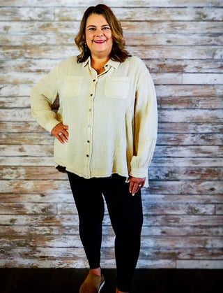 Woman wearing a beige jacket over a white shirt with black pants against a wooden paneled wall.