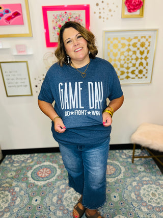 Woman wearing a blue 'GAME DAY GO FIGHT WIN' t-shirt in a room with decorative wall art.