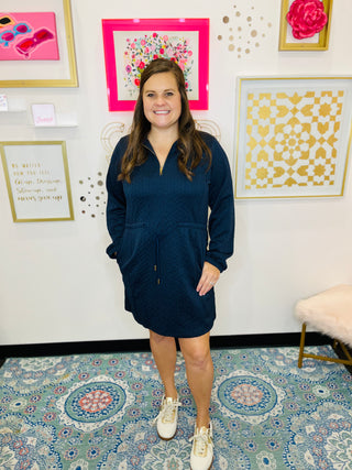 Woman wearing a navy blue dress standing in a room with decorative wall art and patterned carpet.