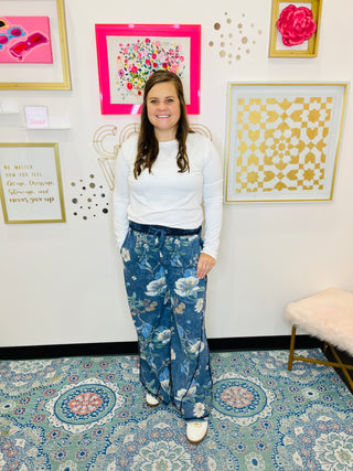 Woman in a white top and blue floral pants standing in a room with decorative wall art.