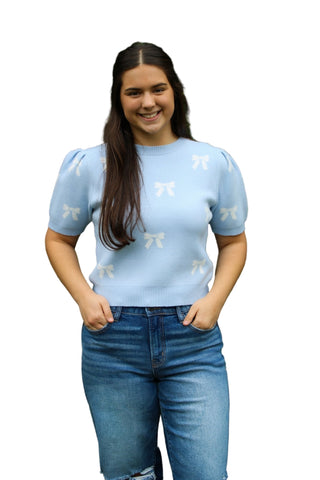 Woman wearing a light blue sweater with white patterns and jeans, standing against a green hedge.